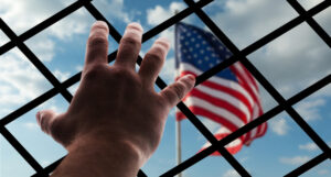 Mano sobre una reja, al fondo la bandera de Estados Unidos